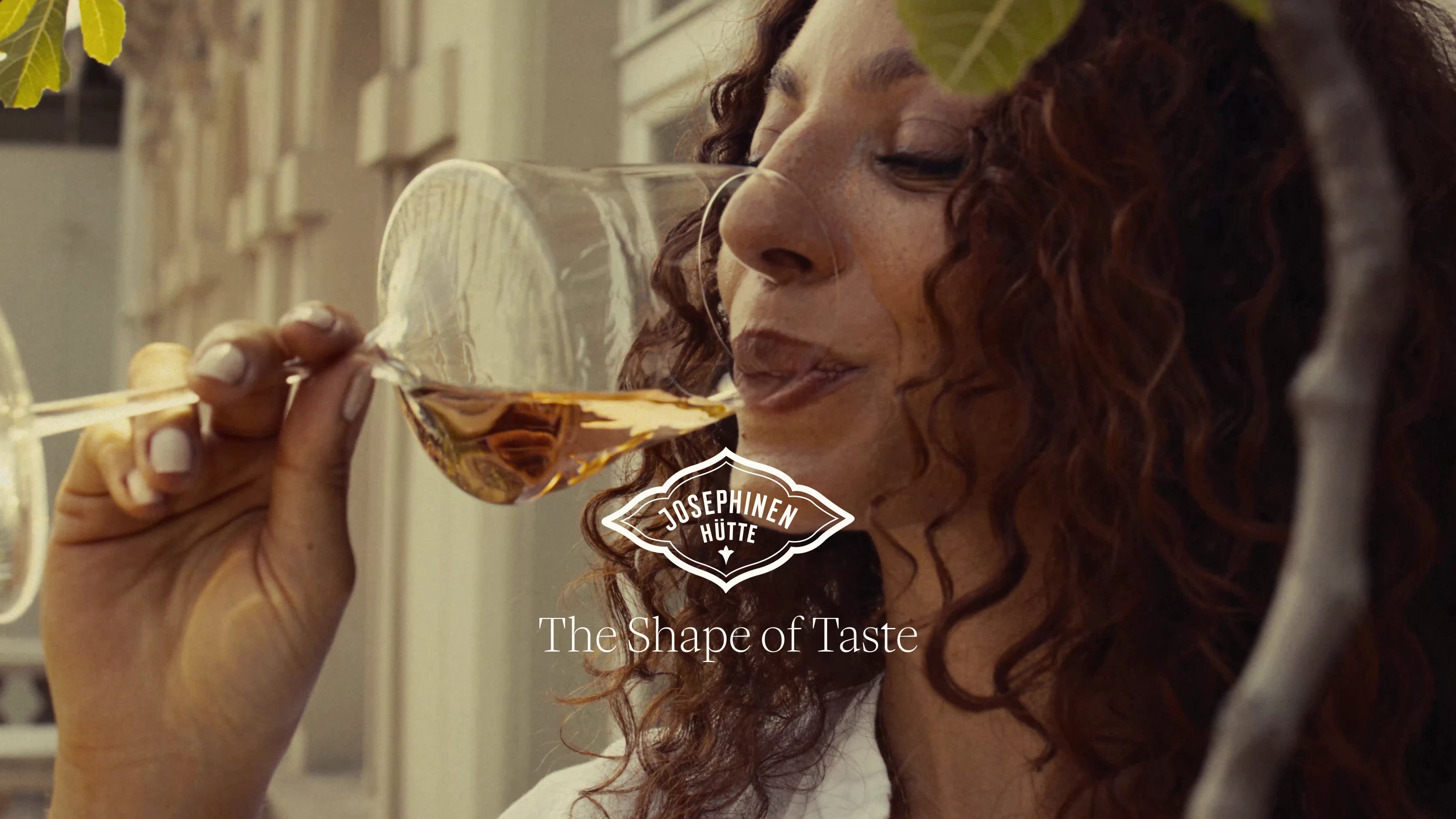 A woman with curly brown hair holds a clear wine glass containing an amber liquid to her lips in an outdoor setting. A white and black logo for Josephinenhütte with the tagline 'The Shape of Taste' is overlaid in the center.
