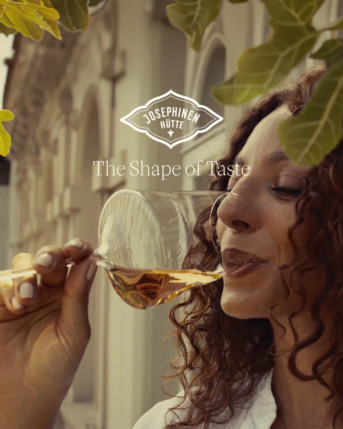 A woman with curly brown hair holds a clear wine glass containing an amber liquid to her lips in an outdoor setting. A white and black logo for Josephinenhütte with the tagline 'The Shape of Taste' is overlaid in the center.